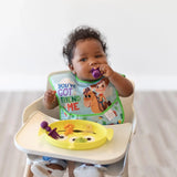A baby in a high chair eagerly chews purple utensils while eating from a Bumkins Silicone Grip Dish: Toy Story Alien.