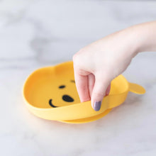 Load image into Gallery viewer, A hand holds a Bumkins Silicone Grip Dish: Winnie The Pooh, demonstrating the suction cup bottom on a marble surface.
