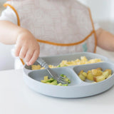Close-up of a child in a beige bib enjoying veggies with a fork from a Bumkins Silicone Grip Dish: Gray at a white table.
