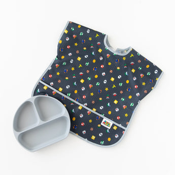 Bumkins Junior Bib: Super Mario™ Lineup lays flat on a white background next to a gray silicone divided plate.