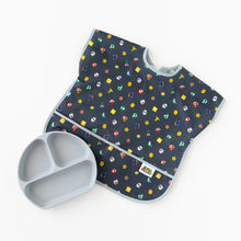 Load image into Gallery viewer, Bumkins Junior Bib: Super Mario™ Lineup lays flat on a white background next to a gray silicone divided plate.
