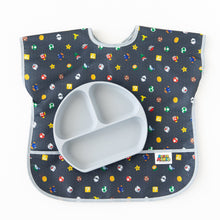 Load image into Gallery viewer, A gray divided plate sits on top of a Bumkins Junior Bib: Super Mario™ Lineup on a white background.
