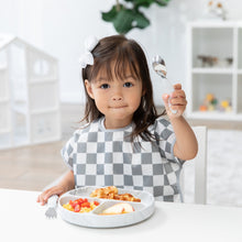 Load image into Gallery viewer, A girl with a bow in a gray &amp; white checkered bib holds Bumkins Spoon + Fork: Marble near a plate with breakfast food on a white table.
