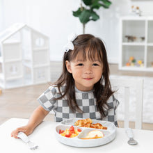 Load image into Gallery viewer, A smiling girl wearing a checkered bib eats breakfast from a Bumkins Silicone Grip Dish: Marble with matching utensils in a bright playroom.

