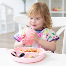 Load image into Gallery viewer, A girl smiles as she drinks from a Bumkins Silicone Straw Cup with Lid: Pink; a pink plate and utensils sit on the table in front of her.
