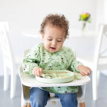 Load image into Gallery viewer, A curly-haired toddler in a high chair wearing a green bib eats from a Bumkins Silicone Grip Dish: Sage with matching utensils.
