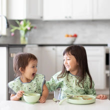 Load image into Gallery viewer, Two kids in matching bibs smile at each other while eating at a white table; one uses a Bumkins Silicone Grip Dish: Sage.
