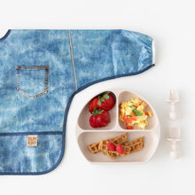 Load image into Gallery viewer, Bumkins Sleeved Bib: Vintage Denim lays flat next to a plate with berries, eggs, &amp; waffles, and toddler utensils on a white background.
