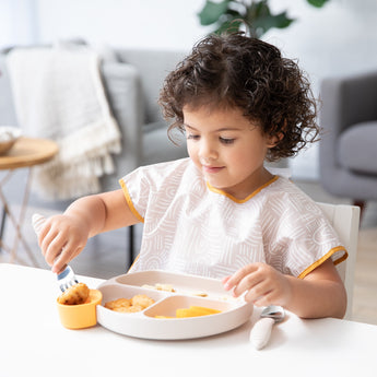 A young child wearing a beige Bumkins Junior Bib eats from a beige dish with beige utensils at a white table.