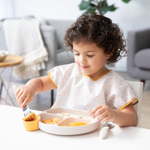 Load image into Gallery viewer, A young child wearing a beige Bumkins Junior Bib eats from a beige dish with beige utensils at a white table.
