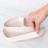 A hand holds a Bumkins Silicone Grip Dish: Sand, demonstrating the suction cup bottom on a marble surface.