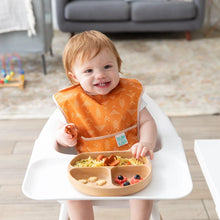 Load image into Gallery viewer, A toddler in a high chair wearing an orange bib enjoys food from Bumkins Silicone Grip Dish: Wood Grain.
