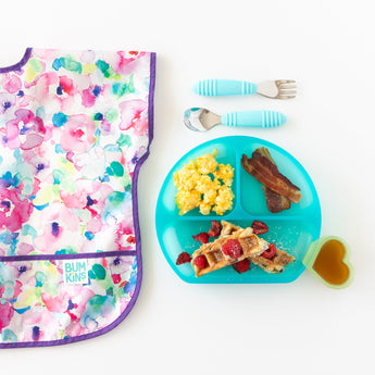 Bumkins Silicone Grip Dish: Blue Jelly with waffles, eggs, and bacon, utensils, and a heart cup sit next to a bib on a white background.