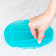 Load image into Gallery viewer, A hand holds a Bumkins Silicone Grip Dish: Blue Jelly, demonstrating the suction cup bottom on a marble surface.
