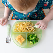 Load image into Gallery viewer, A child with a colorful bib enjoys spaghetti, cornbread, and broccoli from a Bumkins Silicone Grip Dish: Green Jelly at a white table.
