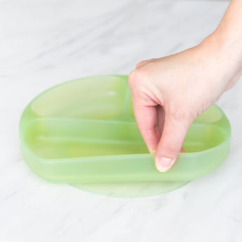 A hand holds a Bumkins Silicone Grip Dish: Green Jelly, demonstrating the suction cup bottom on a marble surface.