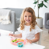A curly-haired girl smiles while eating from a Bumkins Silicone Grip Dish: Pink Jelly at a white table, with a plant and sofa behind her.