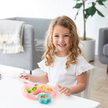 Load image into Gallery viewer, A curly-haired girl smiles while eating from a Bumkins Silicone Grip Dish: Pink Jelly at a white table, with a plant and sofa behind her.
