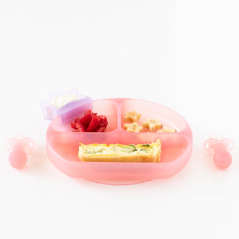 Bumkins Silicone Grip Dish: Pink Jelly, filled with berries, bananas, and quiche, a star cup of yogurt, and utensils on a white background.