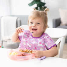 Load image into Gallery viewer, A toddler with pigtails is smiling at a white table, wearing a purple bib and enjoying a meal from a Bumkins Silicone Grip Dish: Pink Jelly.
