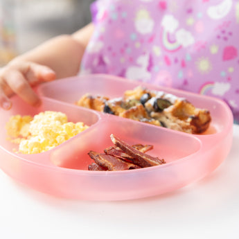 Close-up of a child eating eggs, bacon, & blueberry-topped waffles from a Bumkins Silicone Grip Dish: Pink Jelly.
