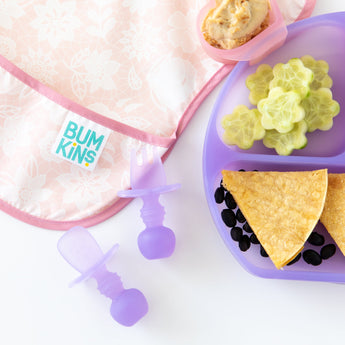 Close-up of a pink bib, Bumkins Silicone Grip Dish: Purple Jelly with tortillas, beans, & cucumber, and toddler utensils on a white background.