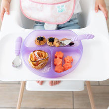 Load image into Gallery viewer, A child in a high chair has a Bumkins Silicone Grip Dish: Purple Jelly with cinnamon rolls, melon flowers, berries, and purple utensils.
