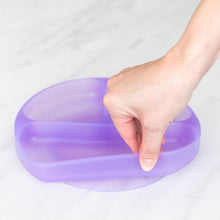 Load image into Gallery viewer, A hand holds a Bumkins Silicone Grip Dish: Purple Jelly, demonstrating the suction cup bottom on a marble surface.
