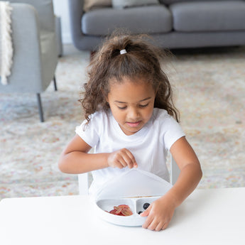 A girl in a white t-shirt removes a Bumkins Silicone Stretch Lid for Grip Dish 3 Section, Grip Plate from a white plate on a white table.