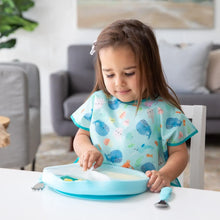 Load image into Gallery viewer, A young child in a blue bib lifts a lid from a Bumkins Silicone Grip Dish with Lid (5 Section): Blue at a white table in a cozy living room.
