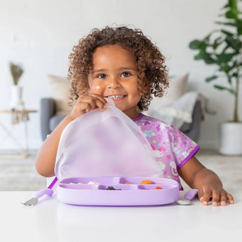 A curly-haired child in a purple bib lifts the lid from a Bumkins Silicone Grip Dish with Lid (5 Section): Lavender at a white table.