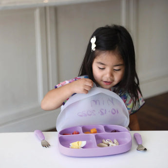 A child with long hair sits at a table, excitedly opening the lid of a Bumkins Silicone Grip Dish with Lid (5 Section): Lavender.