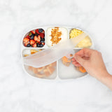 A hand lifts the lid of a Bumkins Silicone Grip Dish with Lid (5 Section): Marble filled with breakfast foods on a marble surface.