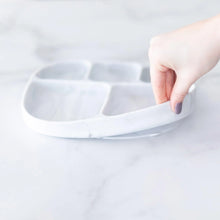 Load image into Gallery viewer, A hand holds a Bumkins Silicone Grip Dish with Lid (5 Section): Marble, demonstrating the suction cup bottom on a marble surface.
