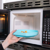 A hand places a Bumkins Silicone Grip Plate: Blue filled with food into an open microwave.
