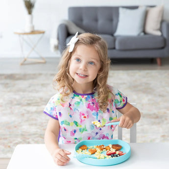A curly-haired young girl in a floral bib smiles while enjoying a meal from a Bumkins Silicone Grip Plate: Blue on a white table.