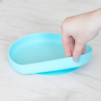 A hand holds a Bumkins Silicone Grip Plate: Blue, demonstrating the suction cup bottom on a marble surface.