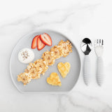 Bumkins Silicone Grip Plate: Gray holds banana, oats, yogurt, berries, & heart-shaped waffles beside gray utensils on a marble background.