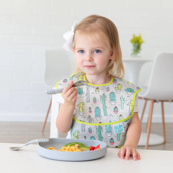 A toddler with a bow happily eats macaroni and strawberries from a Bumkins Silicone Grip Plate: Gray at a white table, wearing a cactus bib.