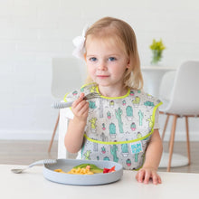 Load image into Gallery viewer, A toddler with a bow happily eats macaroni and strawberries from a Bumkins Silicone Grip Plate: Gray at a white table, wearing a cactus bib.
