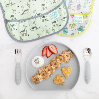 Bumkins Silicone Grip Plate: Gray with waffles, yogurt, strawberries, and granola lays near utensils and two bibs on a marble background.