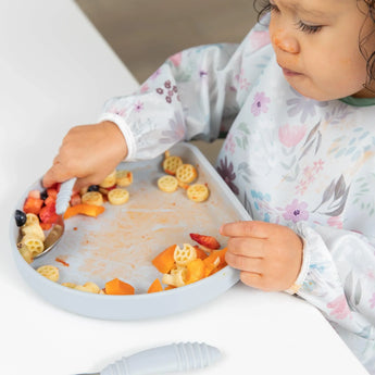 A toddler in a floral bib enjoys pasta and veggies with a spoon from a Bumkins Silicone Grip Plate: Gray on a white table.