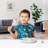 A smiling child in a colorful bib holds a fork, with scrambled eggs on Bumkins Silicone Grip Plate: Gray in front of them on a white table.