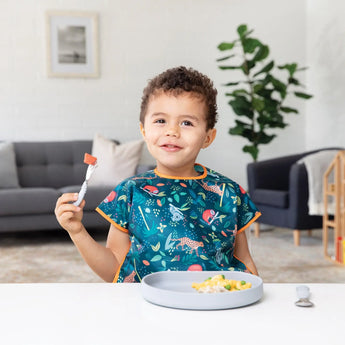 A smiling child in a colorful bib holds a fork, with scrambled eggs on Bumkins Silicone Grip Plate: Gray in front of them on a white table.