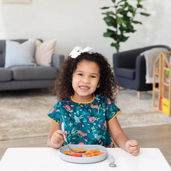 A smiling girl with curly hair enjoys chicken nuggets from a Bumkins Silicone Grip Plate: Gray at a white table in a cozy living room.