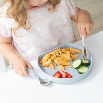 A child in a pink bib enjoys pasta, cucumber, & strawberries with utensils from a Bumkins Silicone Grip Plate: Gray at a white table.