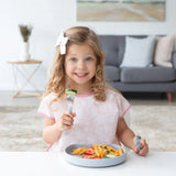 A curly-haired girl smiles, holding a fork over a Bumkins Silicone Grip Plate: Gray filled with pasta on a white table.