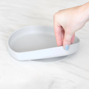 A hand holds a Bumkins Silicone Grip Plate: Gray, demonstrating the suction cup bottom on a marble surface.