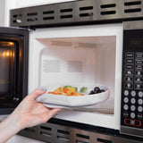 A hand places a Bumkins Silicone Grip Plate: Marble filled with food into an open microwave.