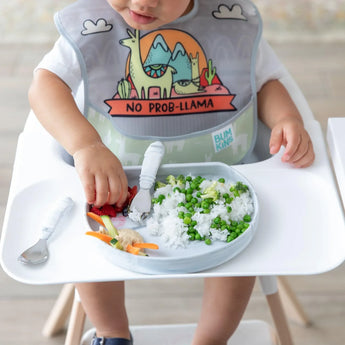 A baby wearing a llama bib in a high chair enjoys rice with peas from a Bumkins Silicone Grip Plate: Marble.